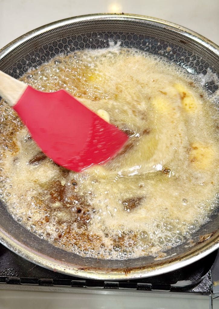 A red spatula stirs crispy bacon bits in a hot pan of bubbling oil and foam on a stove. The motion creates a ripple effect across the surface of the liquid. The pan has a non-stick, textured bottom.