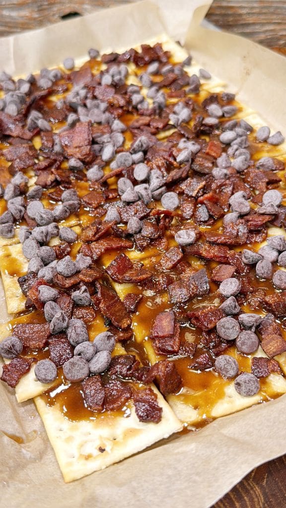 Rectangular pastry topped with evenly scattered bacon bits and chocolate chips on a brown parchment paper-lined tray. The pastry appears to be glazed with a caramel-like sauce.