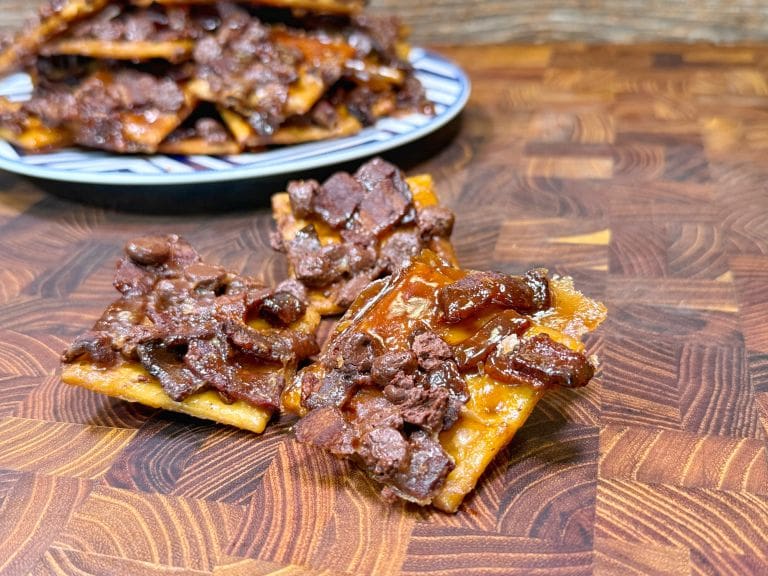 Close-up of crispy toffee bark topped with melted chocolate and nuts, resting on a wooden surface. A plate with a pile of more bark pieces is blurred in the background.