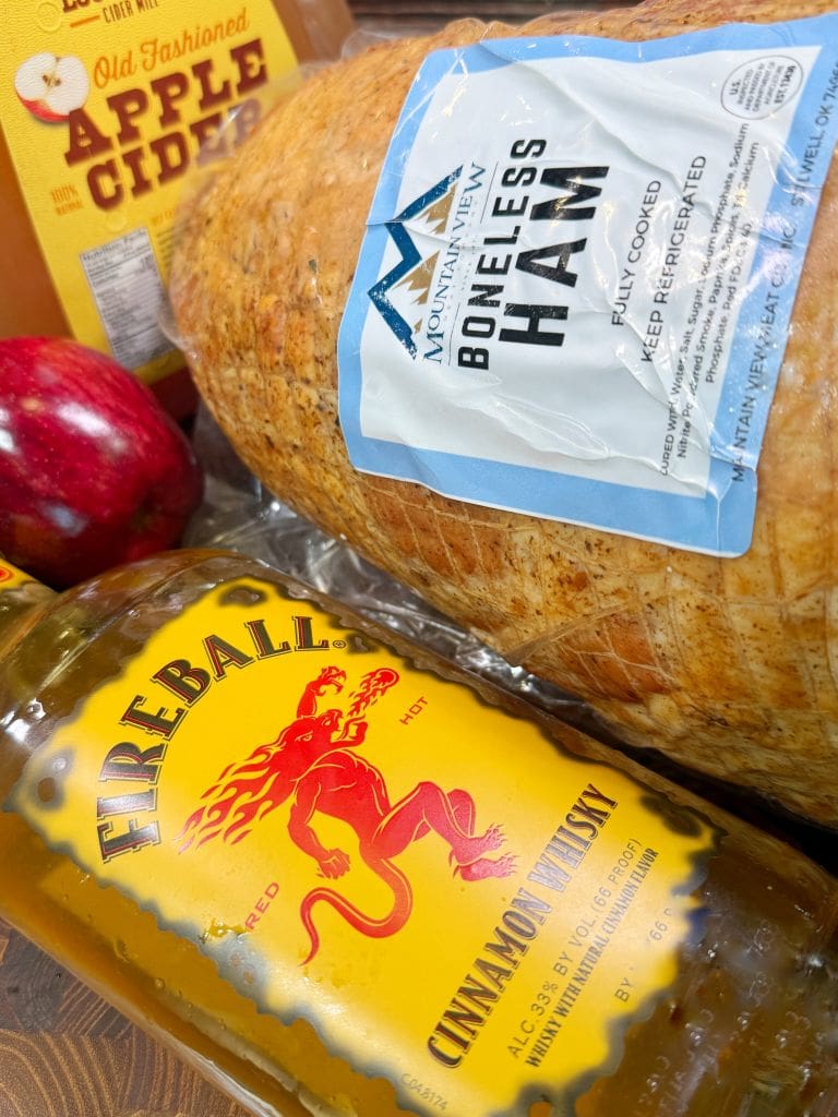 Bottles of apple cider and Fireball Cinnamon Whisky are on a table beside a large packaged boneless ham and a red apple. The hams label reads Mountain Valley Boneless Ham. The scene is set on a wooden surface.