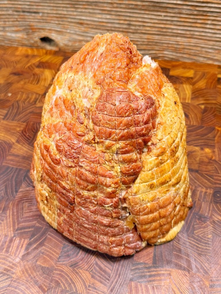 A whole, cooked ham with a browned, crispy exterior, sitting on a wooden cutting board with a textured grain pattern. The hams surface is marked with a crisscross pattern from the cooking process.