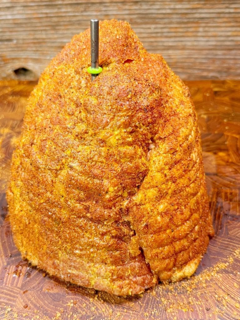 A seasoned, roasted ham stands upright on a wooden cutting board. A metal meat thermometer is inserted into the top, surrounded by a textured, golden-brown crust. The background features a rustic wooden wall.