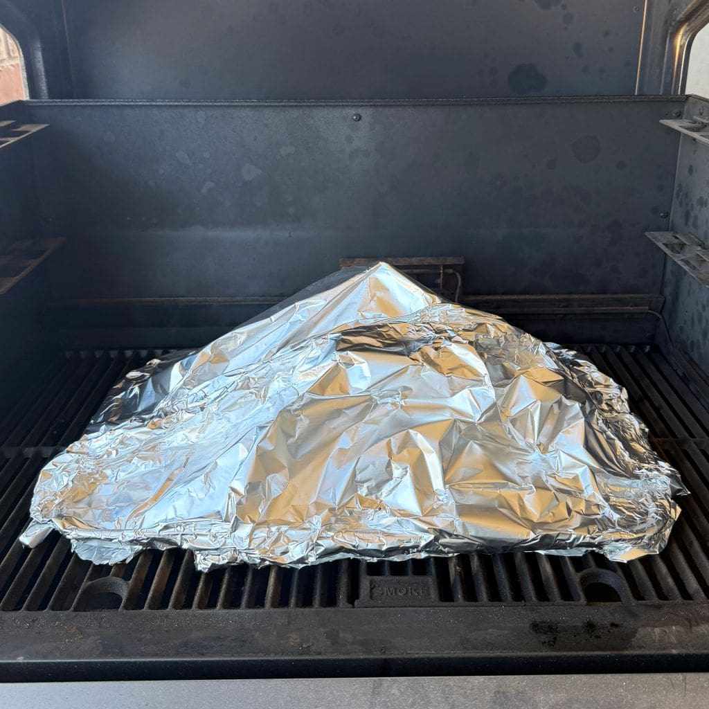 A large piece of food is covered with crumpled aluminum foil on a barbecue grill. The grill lid is open, and the interior is slightly dark with visible grill marks.