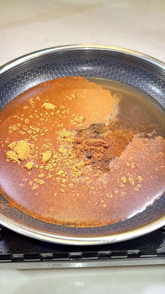 A non-stick pan on a stovetop is filled with water and a mixture of spices, including visible brown and yellow powders, forming a blended pattern as they dissolve.