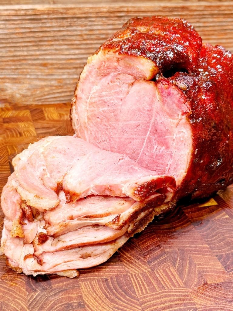 A glazed, cooked ham sits on a wooden cutting board. Several slices are neatly cut and fanned out in front, revealing a juicy, pink interior. The ham has a slightly crispy, caramelized crust on the outside.  This is a delicious smoked ham recipe