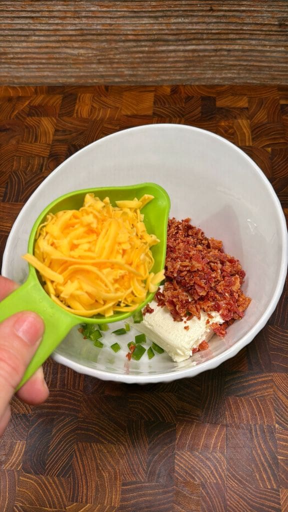 A green measuring cup holds shredded cheddar cheese above a white bowl containing chopped bacon, cream cheese, and green herbs. The bowl is placed on a wooden surface.