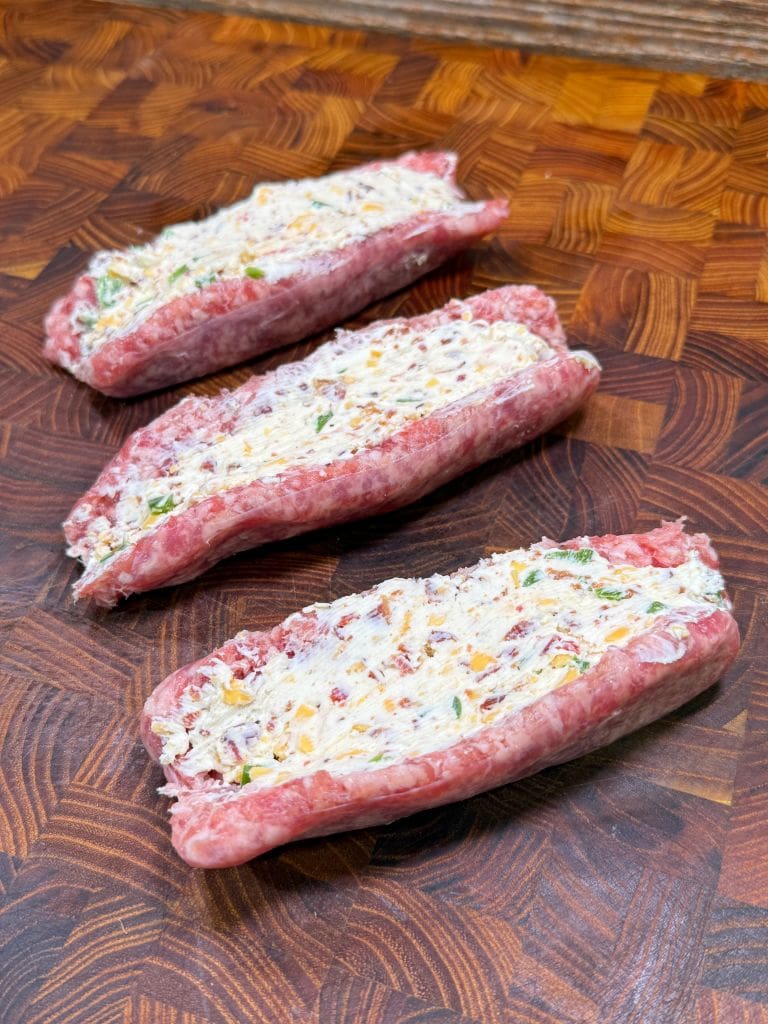 Three raw, partially sliced beef rolls stuffed with a mixture of cream cheese, chopped herbs, and diced vegetables are arranged on a wooden cutting board with a geometric pattern. Picture of jalapeno popper stuffed bratwurst being ready to be cooked.