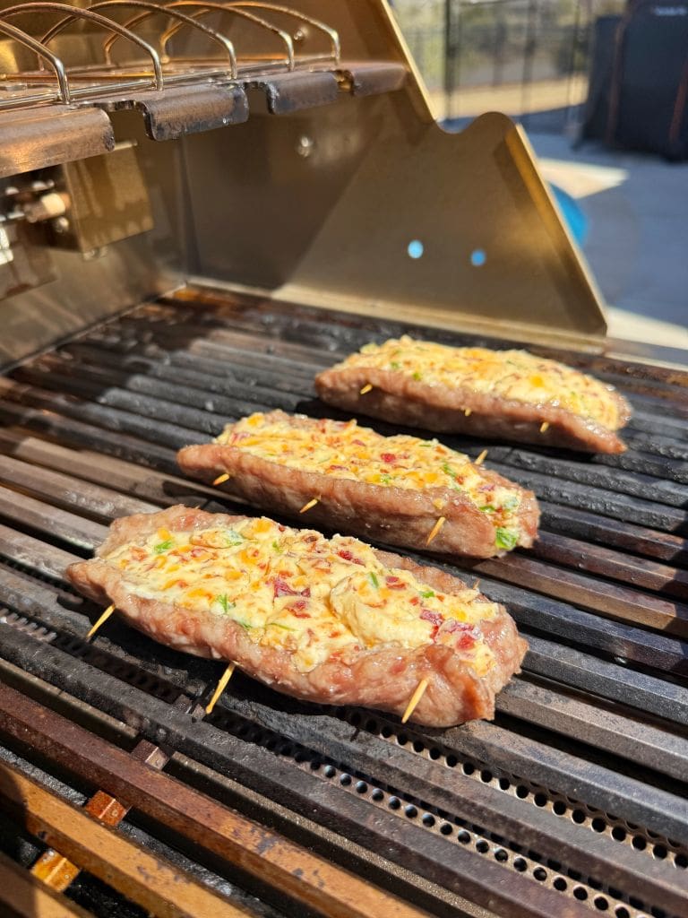 Three stuffed steaks are grilling on a barbecue grill. The steaks are topped with a mixture of cheese, herbs, and spices, and are held together with wooden skewers. The grill is outdoors, and the sun is shining brightly.