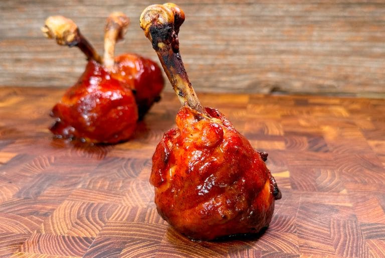 Close-up of two grilled chicken drumsticks coated with a glossy barbecue sauce, placed upright on a textured wooden surface. The background is a warm, rustic wood pattern.