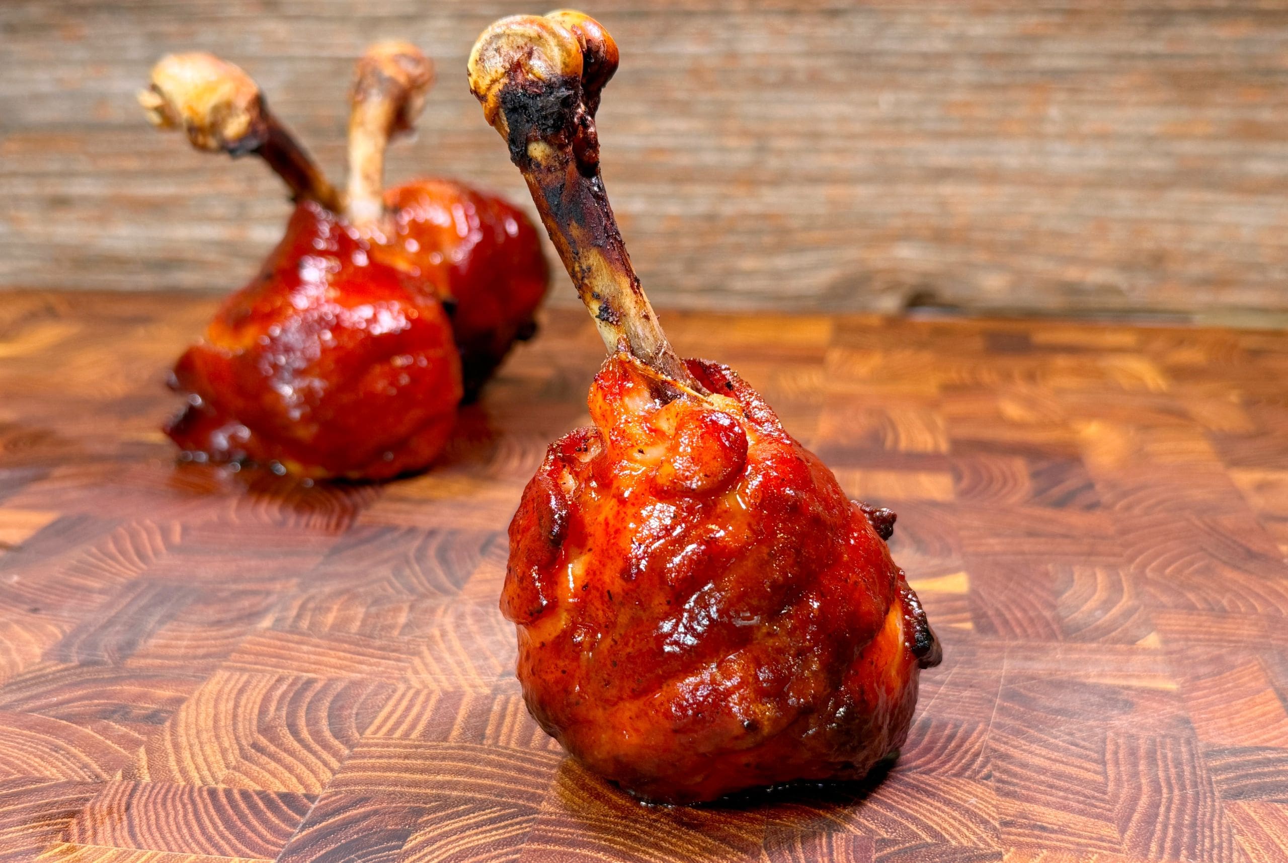 Close-up of two grilled chicken drumsticks coated with a glossy barbecue sauce, placed upright on a textured wooden surface. The background is a warm, rustic wood pattern.