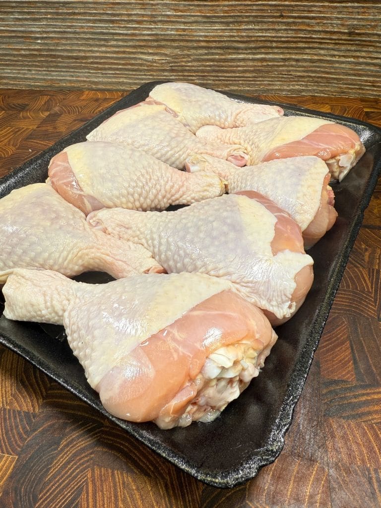 Tray of raw chicken drumsticks and thighs with pale skin, arranged neatly on a dark platter. The wooden surface beneath has a warm, textured appearance.