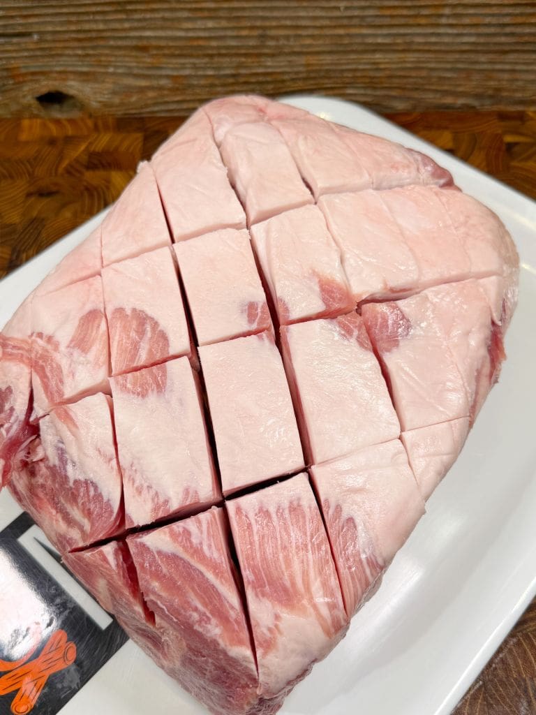 A piece of raw, marbled pork with a criss-cross scoring pattern on the surface, placed on a white rectangular plate. The background shows a wooden texture, adding warmth to the scene.