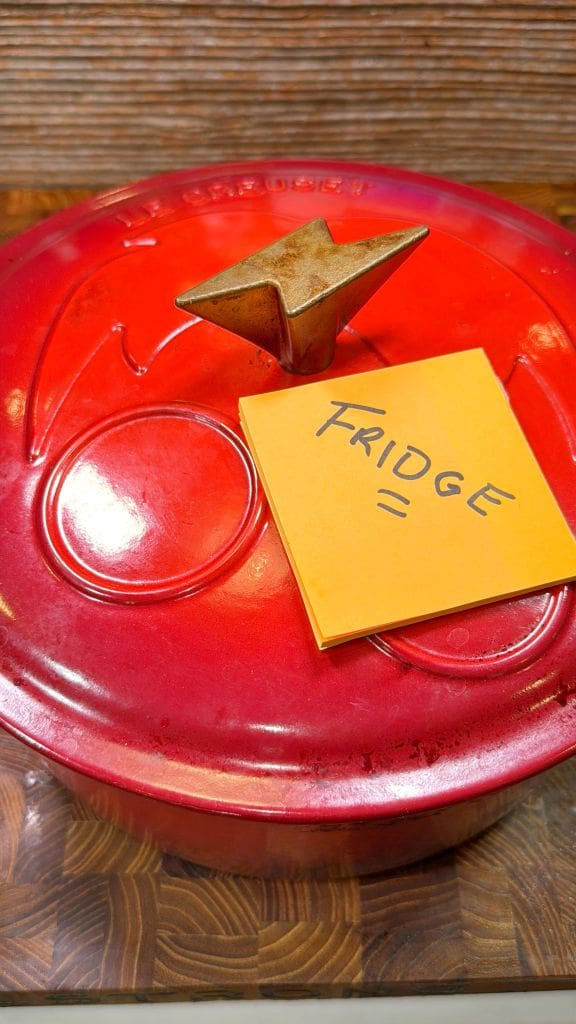 A red pot with a metal handle on a wooden surface. The pot has a sticky note on the lid with the word FRIDGE written on it.