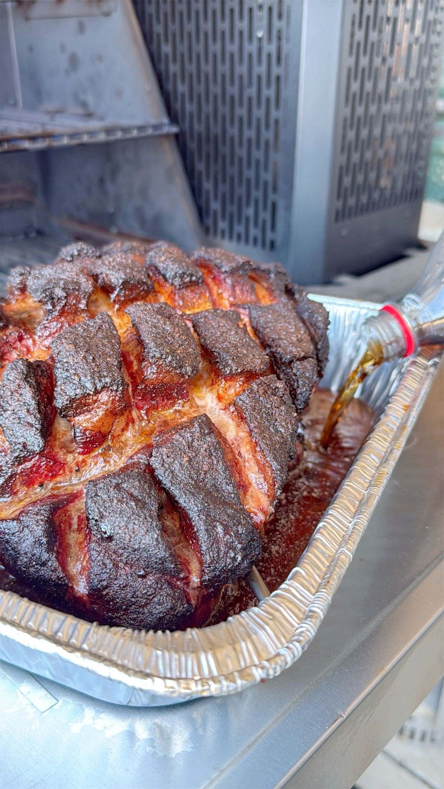 Smoked pork shoulder with a dark, crispy bark in a foil tray, being basted with a liquid poured from a bottle. The meat has a crosshatch pattern. A grill is visible in the background.