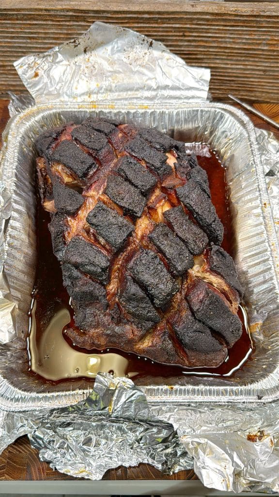 A roasted meat dish in a foil tray, featuring a charred, crisscross pattern on the surface. The meat is resting in juices, with aluminum foil surrounding the tray.