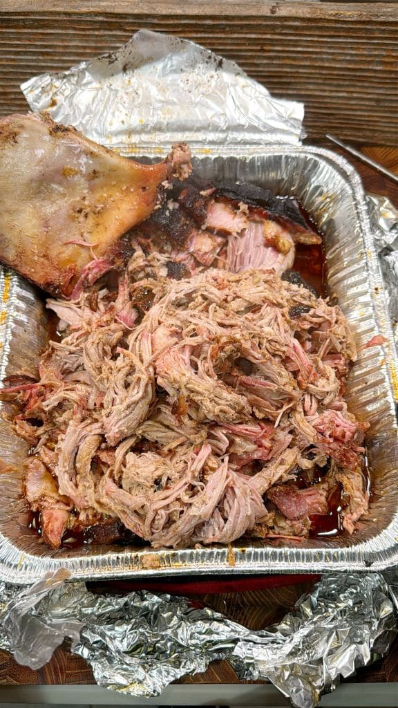 A foil tray filled with smoked pulled pork, showing shredded pieces and a slice of bark. Some foil is crumpled around the tray, and the wood surface underneath is partially visible. The pork is juicy and has a pink, smoky appearance. This recipe is for hot and fast pulled pork
