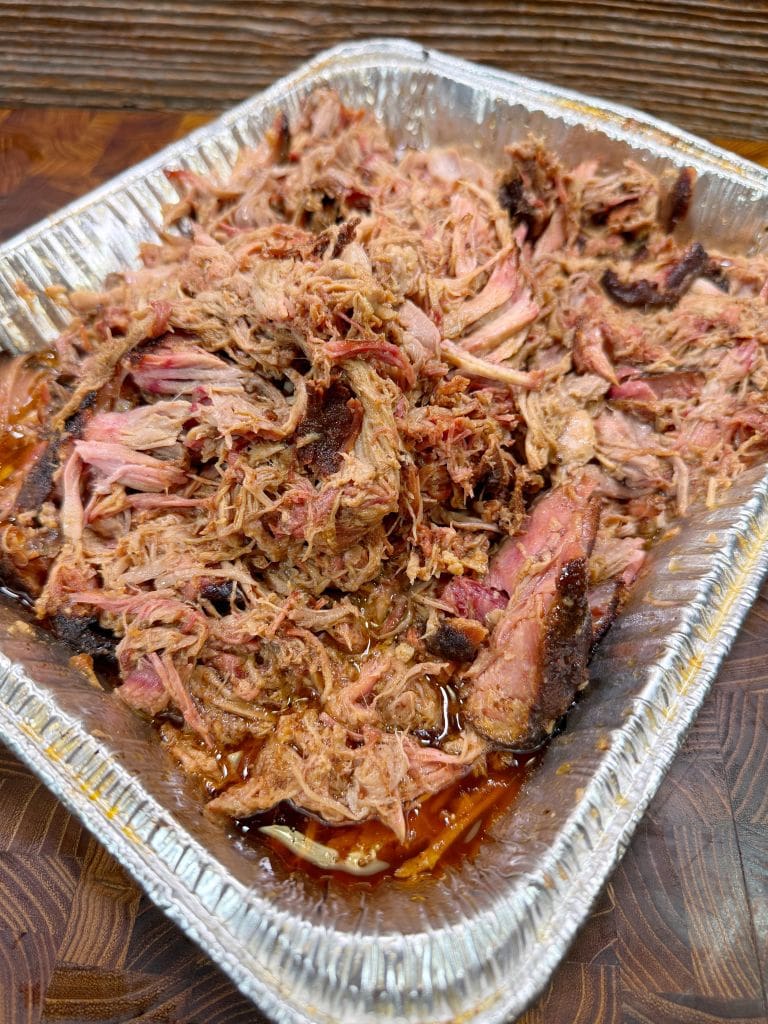 A tray of shredded, smoked pork barbecue, with a mix of well-cooked and pinkish meat, is served in a rectangular aluminum foil container. The meat appears juicy and tender, resting on a wooden surface.