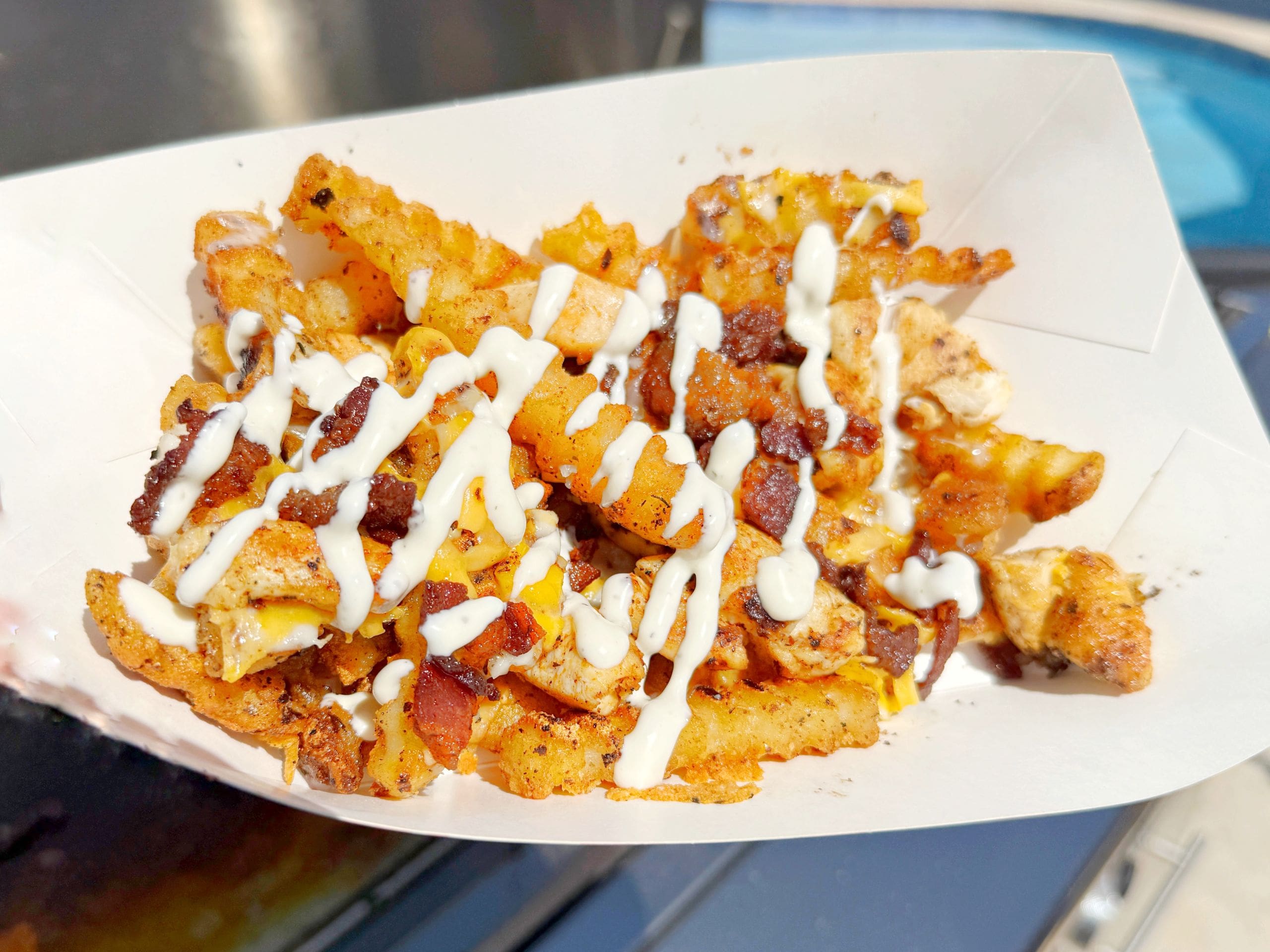 Crinkle-cut fries topped with melted cheddar cheese, chunks of grilled chicken, bits of bacon, and drizzled with ranch dressing, served in a white paper tray.