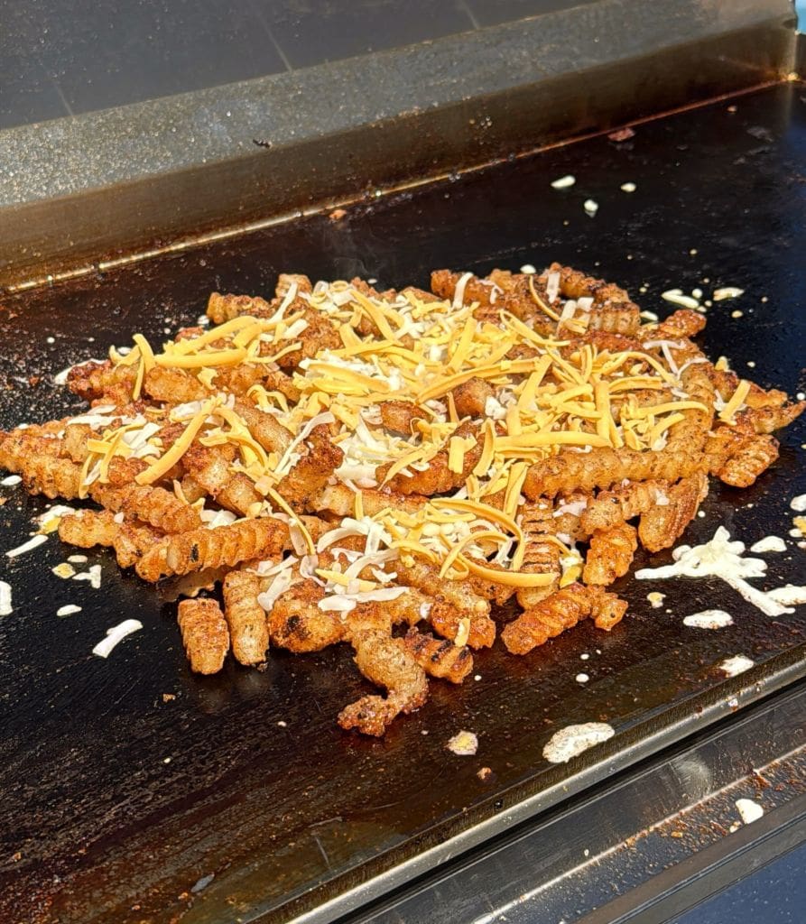 A pile of golden-brown curly fries topped with melted shredded cheese on a flat-top grill. The fries appear crispy and are generously coated with cheddar and mozzarella cheese, glistening under the sunlight.