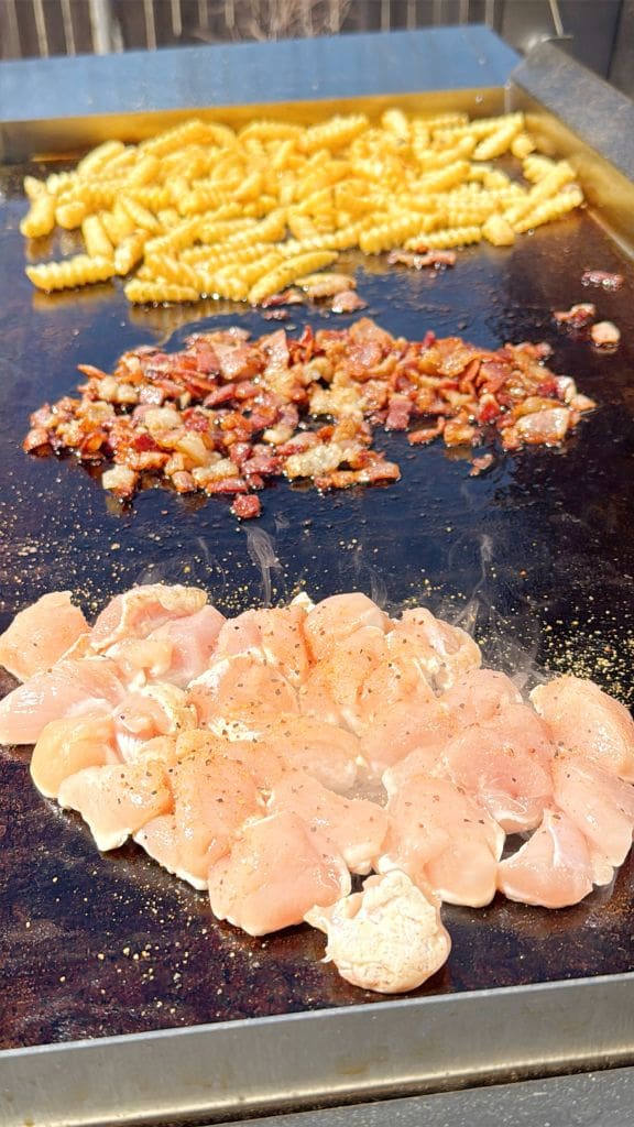 Chicken pieces, diced bacon, and crinkle-cut fries cooking on a large grill. The raw chicken is in the foreground, the crispy bacon in the middle, and the golden fries in the background. Seasoning is sprinkled around the chicken.
