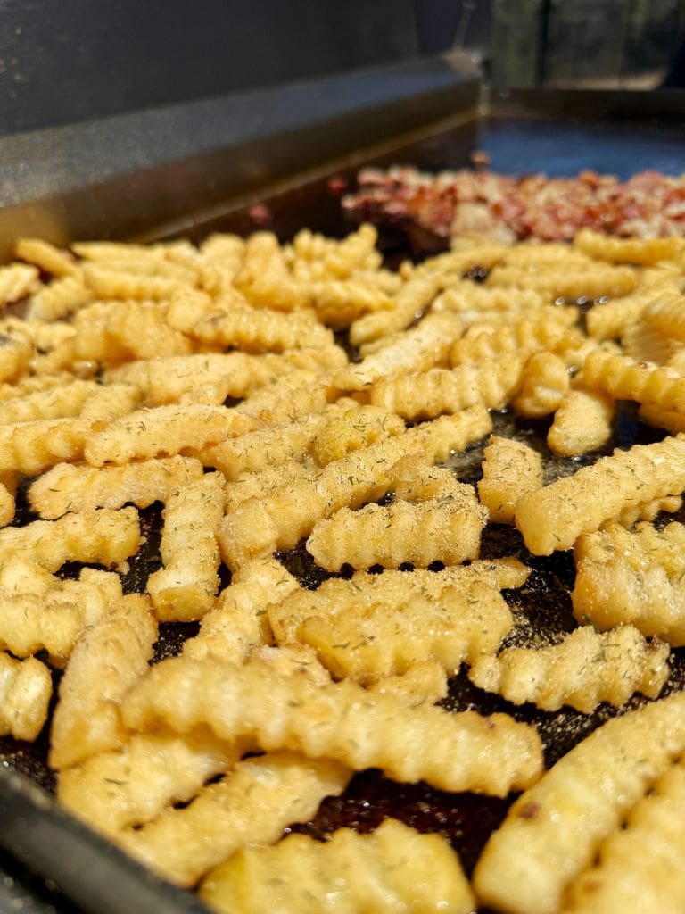 Crinkle-cut fries are sizzling on a hot grill, with some bacon cooking in the background. The fries are golden and slightly crispy, occupying most of the foreground.