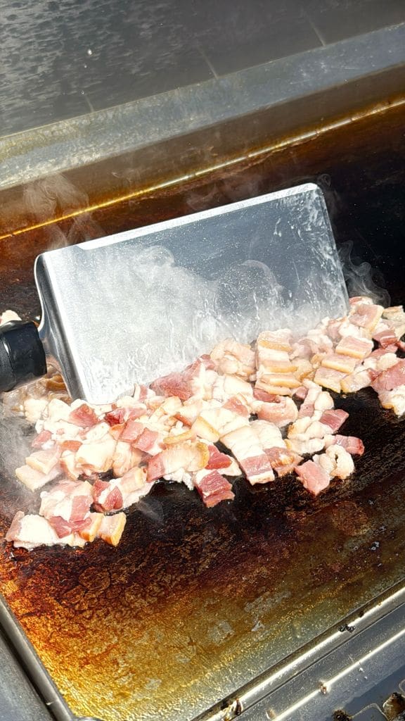 A metal spatula stirring sizzling chunks of bacon on a hot griddle, with steam rising from the cooking meat.