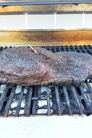 A large, well-seasoned brisket with a dark, crispy crust is cooking on a grill. There is a meat thermometer inserted into the brisket, and the background shows a slightly open grill lid.