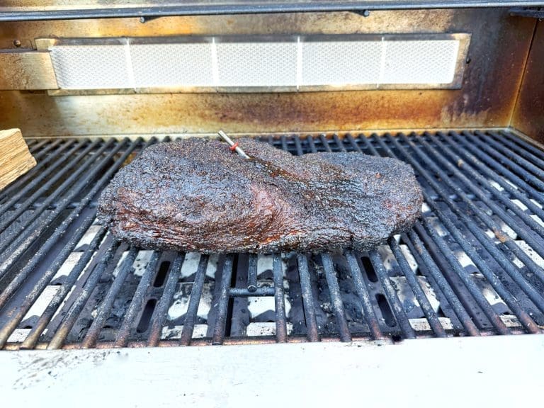 A large, well-seasoned brisket with a dark, crispy crust is cooking on a grill. There is a meat thermometer inserted into the brisket, and the background shows a slightly open grill lid.