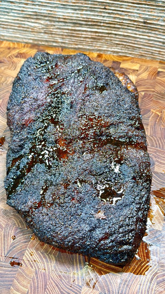 Close-up of a perfectly smoked brisket with a dark, crispy bark glistening under light. The wood-textured cutting board complements the rich, rustic appearance of the brisket.