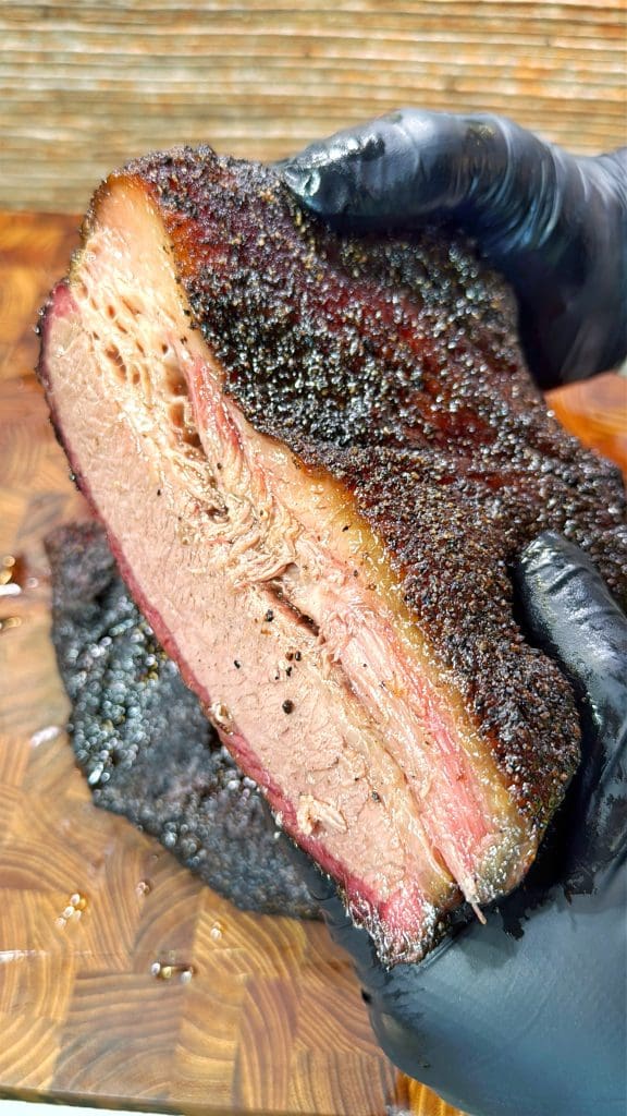 Close-up of a person wearing black gloves holding a thick slice of juicy, seasoned brisket with a dark crust and pink smoke ring. The brisket rests on a wooden surface, showing its tender, marbled texture.