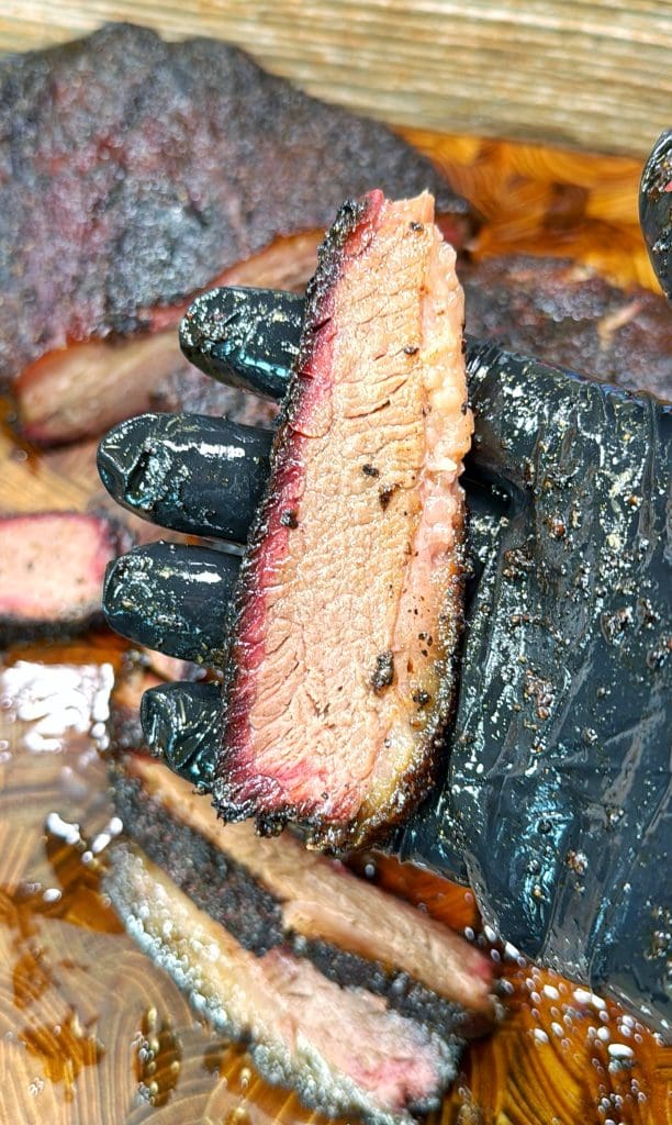 A gloved hand holds a slice of perfectly cooked brisket with a dark, seasoned bark and a visible smoke ring. More slices lie on a wooden cutting board, with juices glistening on the surface.