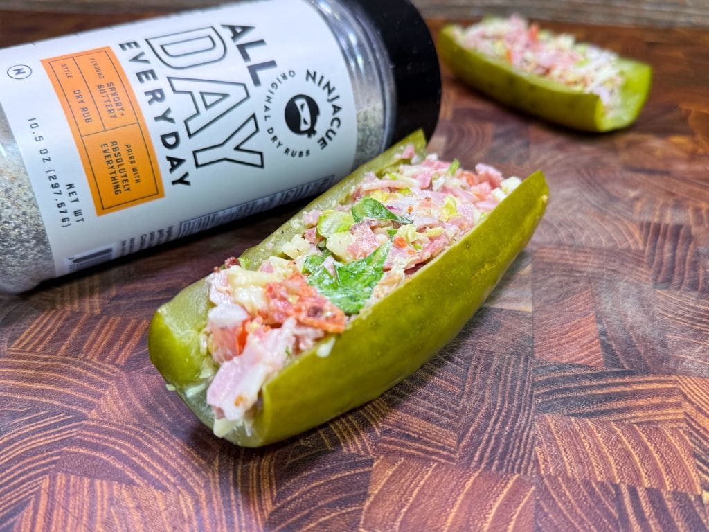 A pickle half filled with a colorful salad mix is placed on a wooden surface. A container labeled All Day Every Day seasoning is lying beside it. The salad appears to include lettuce, tomatoes, and other chopped ingredients.