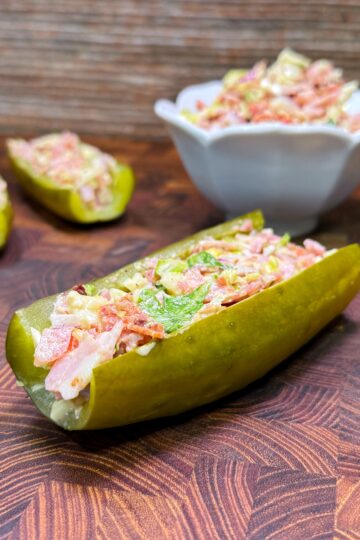 A pickle is sliced lengthwise, filled with a colorful mixture of diced vegetables. More filled pickle halves and a bowl with additional filling are in the background, all placed on a wooden surface.