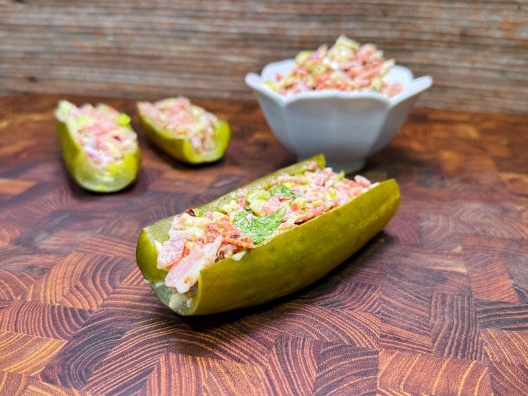 A pickle is sliced lengthwise, filled with a colorful mixture of diced vegetables. More filled pickle halves and a bowl with additional filling are in the background, all placed on a wooden surface.