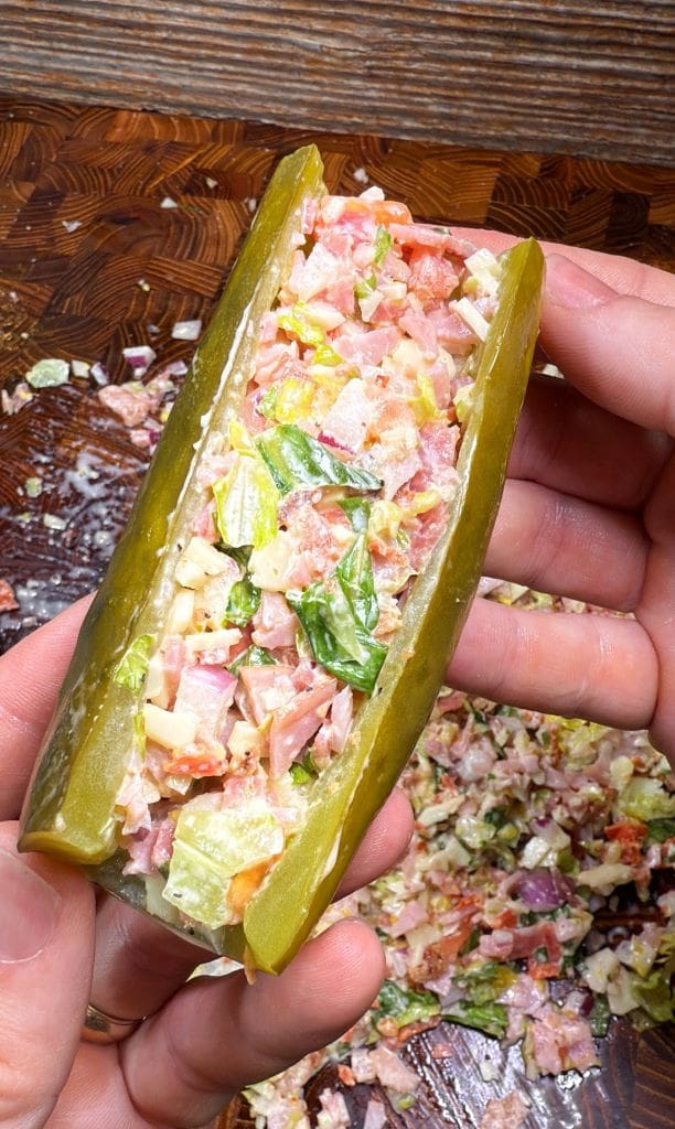 Close-up of a person holding a pickle stuffed with chopped italian sandwich ingredients, including lettuce, tomatoes, onions, and meats. Finely diced bits are visible, and the background shows a wooden surface scattered with similar pieces.