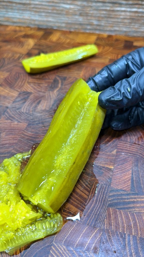 A hand in a black glove holds a sliced pickle on a wooden cutting board. Another pickle slice and some pickle seeds are nearby. The board has a distinctive wood pattern.