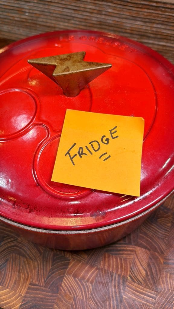 A red pot with a star-shaped handle on the lid is topped with an orange sticky note reading FRIDGE in black letters. The pot rests on a patterned wooden surface.