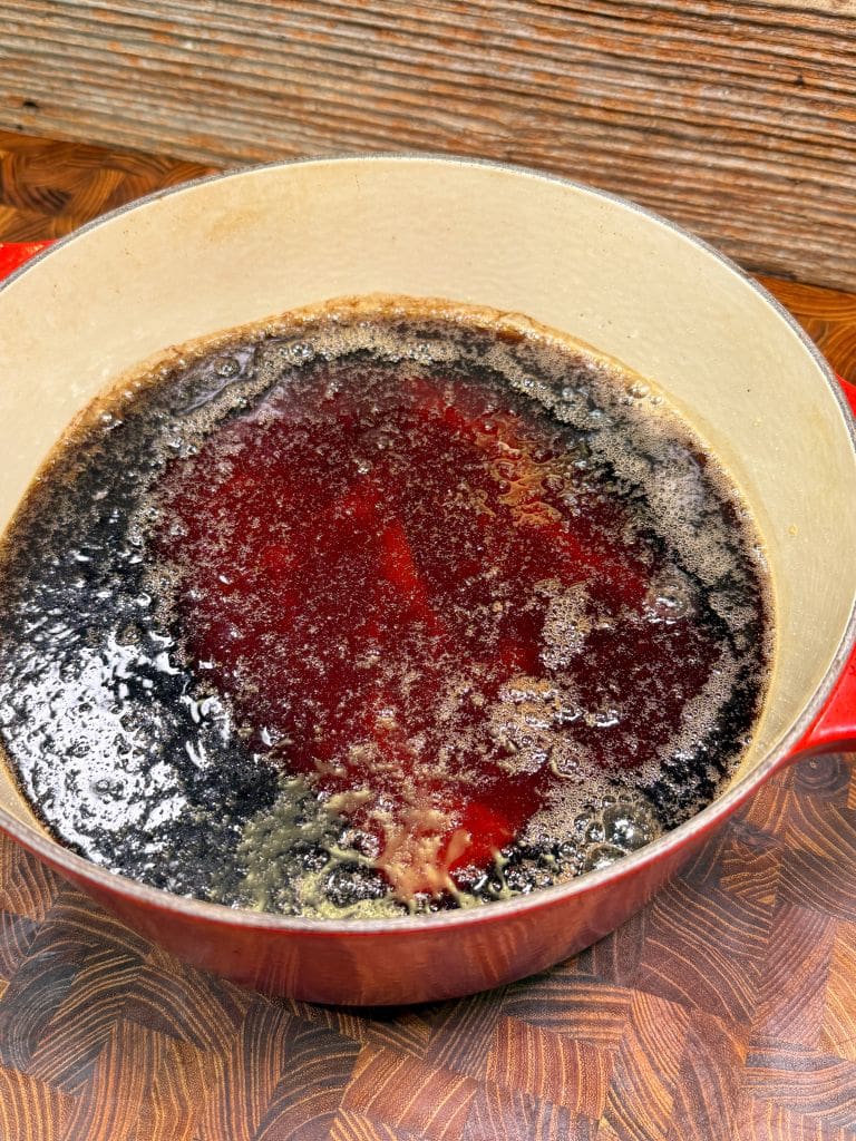 A red pot filled with a reddish-brown liquid, likely cooking sauce or stew, showing a layer of foam on top. The pot is on a wooden surface, and the background is a textured wooden wall.