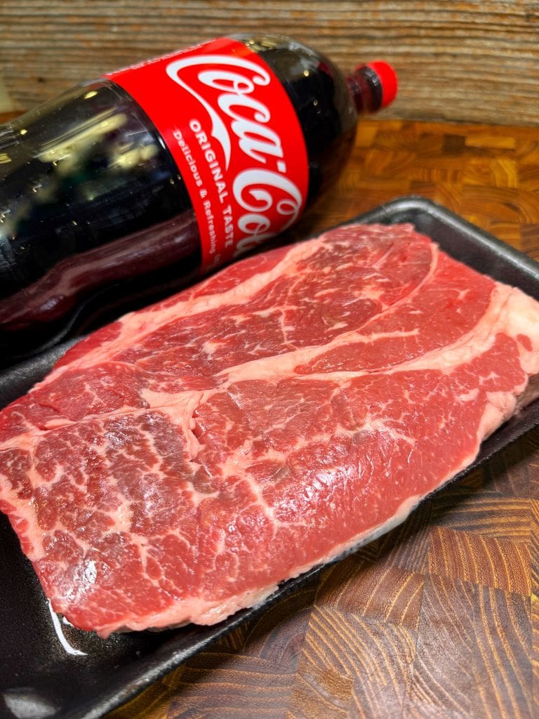 Raw marbled beef steak on a black tray sits on a wooden surface. A bottle of Coca-Cola is placed next to it, angled slightly. The setting suggests preparation for cooking or grilling.