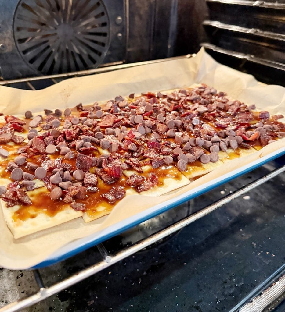 A baking sheet in the oven holds pastry covered with chocolate chips and chopped bacon, placed on parchment paper. The oven rack and interior are visible, and the pastry is baking.