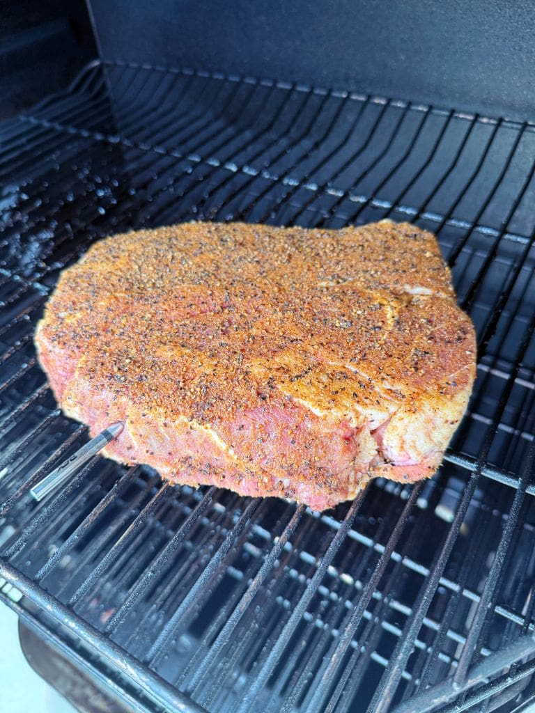 A seasoned cut of meat is on a rack inside a smoker. A thermometer probe is inserted into the meat, which is coated with an even layer of spices and herbs. The scene captures the preparation stage of smoking the meat.