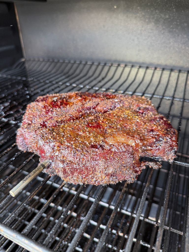 A thick, juicy beef brisket with a dark, seasoned crust is cooking on a metal rack inside a smoker. A temperature probe is inserted into the meat, indicating its being slow-cooked or smoked.