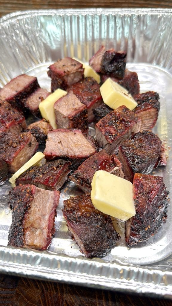 Chunks of grilled meat with a charred crust and succulent interior are topped with slices of butter, all served in a silver tray.