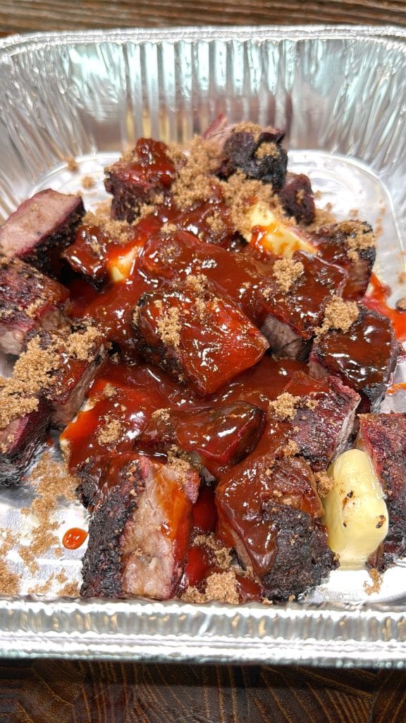 A foil tray filled with smoked beef burnt ends, drizzled with barbecue sauce and sprinkled with brown sugar. The meat is charred on the edges, glistening with sauce, and nestled on pieces of butter.