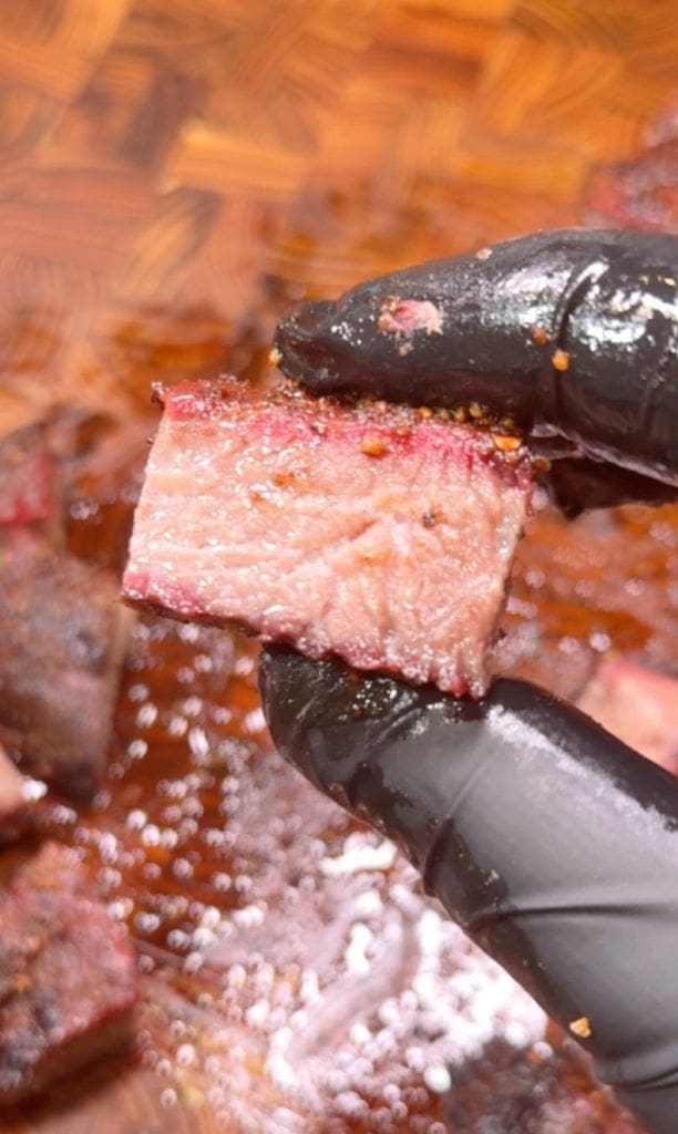 A hand in a black glove holds a thick, juicy slice of seasoned meat, showing a slightly charred edge and a pink interior. This is midway through the cook of chuck roast burnt ends. The background has more pieces of meat on a wooden surface.