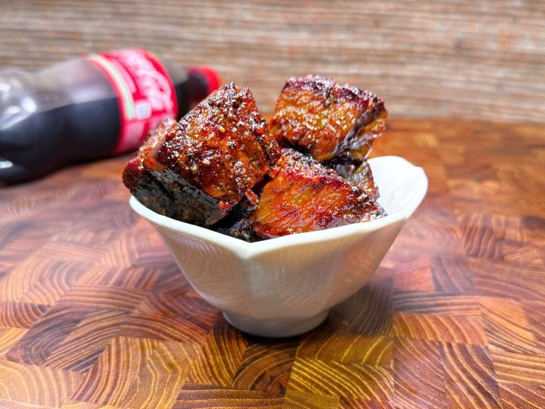 A small white bowl filled with glazed, juicy chunks of meat sits on a patterned wooden surface. A bottle of Coca-Cola lies in the blurred background. The meat is richly colored and glistens, suggesting it is well-seasoned and cooked.