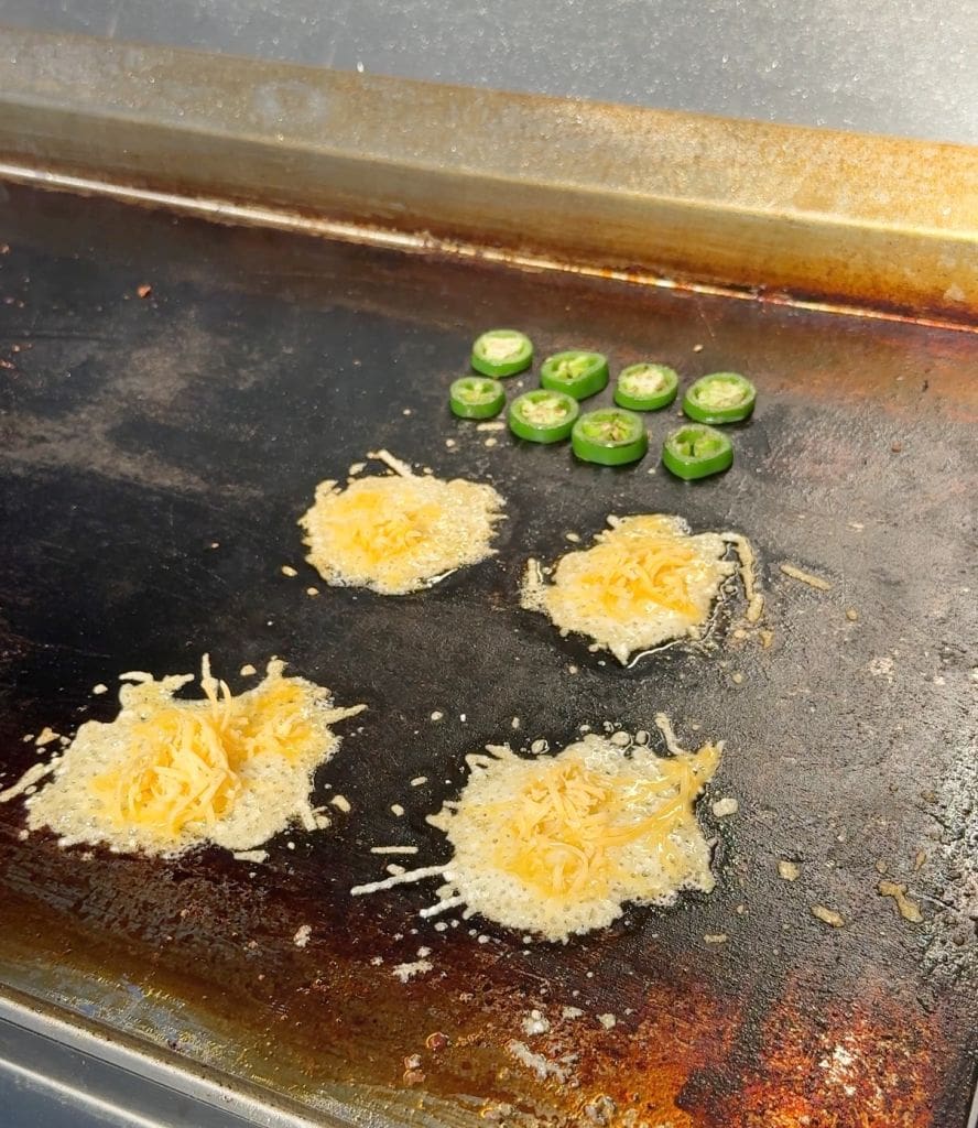 Cheese melting into crispy rounds on a flat grill, with slices of green jalapeños arranged nearby. The grill surface shows areas of browning and seasoning.