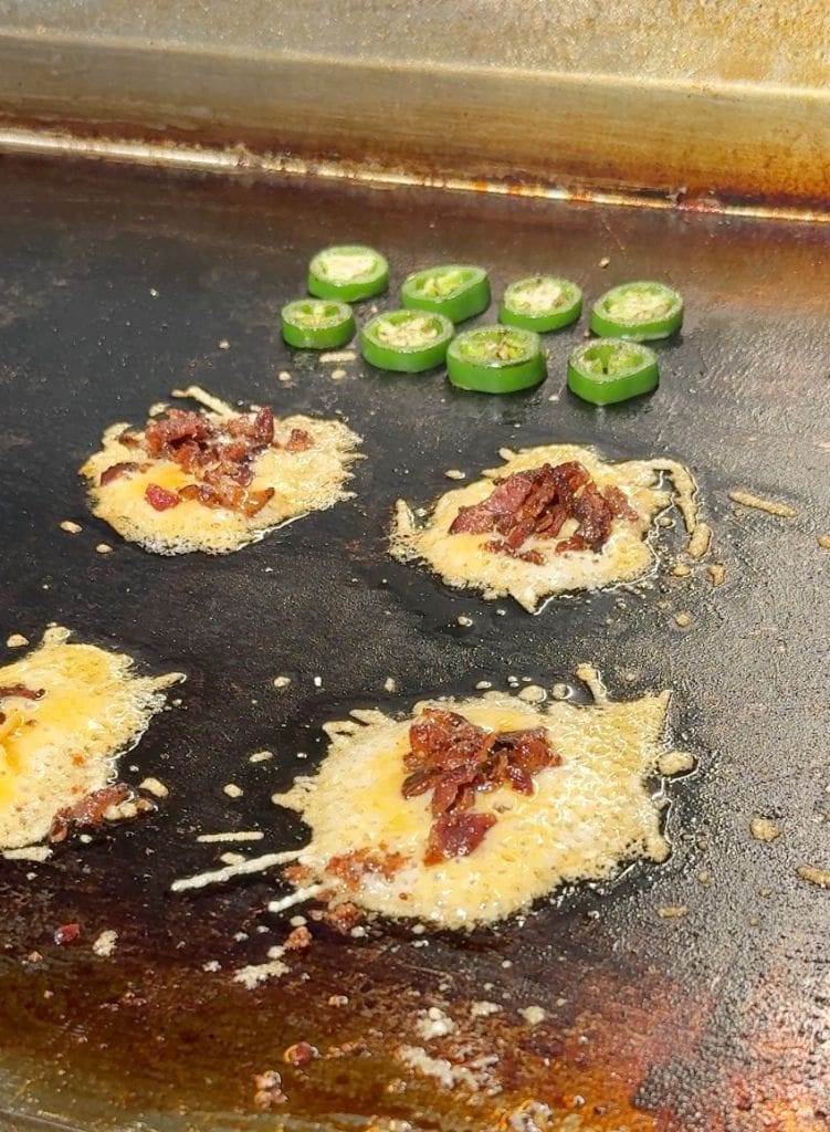 Cheese with bacon bits melting on a griddle, alongside sliced jalapeños. These are parmesan cheese crisps