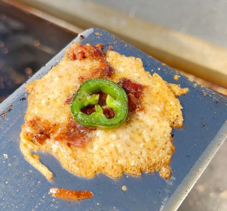 Close-up of a sizzling, crispy cheese slice topped with bacon bits and a single jalapeño slice, resting on a spatula. The cheese is melted and golden brown, with the jalapeño adding contrasting color.