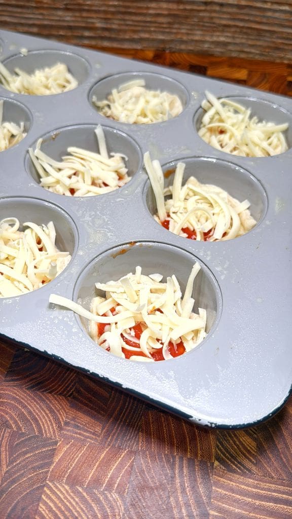 Muffin tin filled with unbaked miniature pizzas. Each cup contains dough topped with tomato sauce and shredded cheese. Wooden surface beneath the tin.